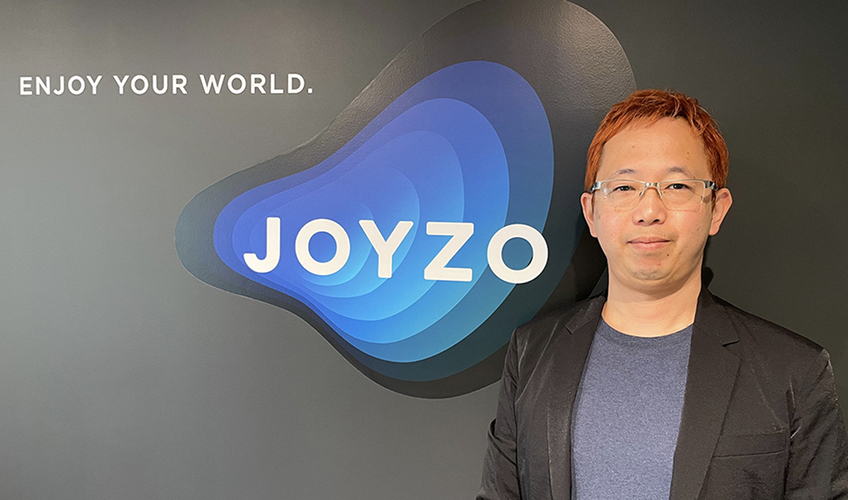 グレーの背景に青いグラデーションのJOYZOロゴと『ENJOY YOUR WORLD.』のキャッチフレーズが配置された企業ブランディング背景の前で、眼鏡をかけた男性が黒いジャケットとグレーのシャツを着用して立っているポートレート写真